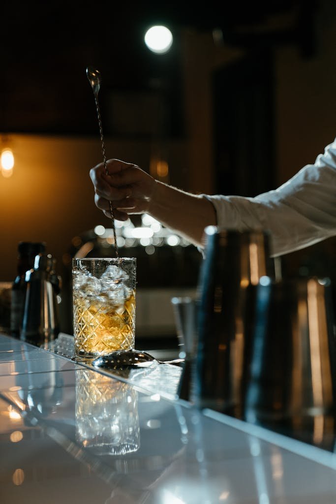 A bartender stirring a whiskey cocktail with a spoon in a dimly lit bar setting.